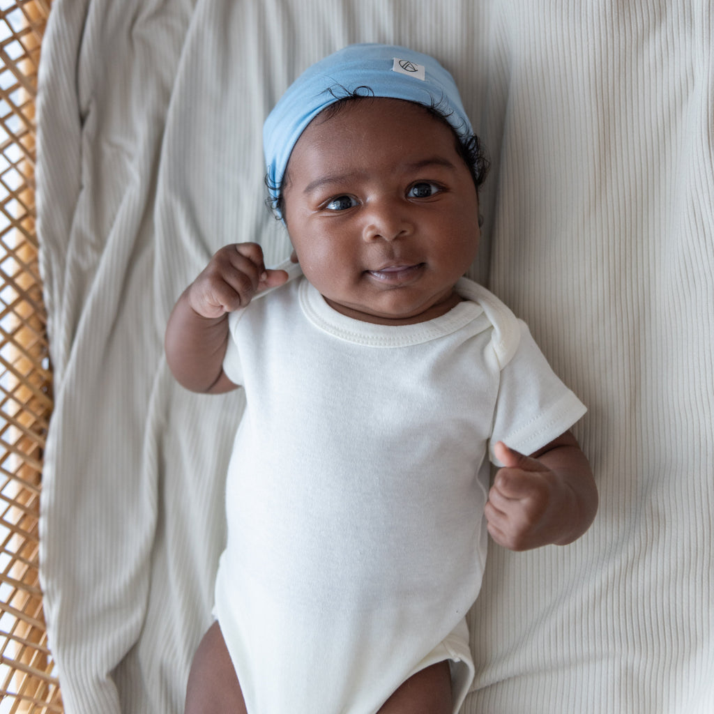 Baby in a white onesie and blue beanie lying on a textured surface